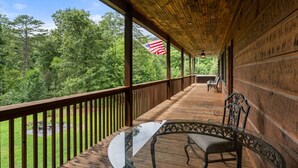 A wooden deck with a glass table and chairs, overlooking a forested area with an American flag.