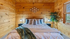 A wooden cabin bedroom with a bed, bedside lamps, a decorative wall piece, and a window with blinds.