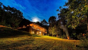 A large, modern house with a spacious deck and a well-lit interior, surrounded by a grassy landscape and trees.