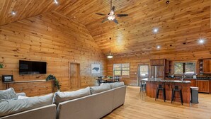 A spacious log cabin interior with a wooden ceiling, a large sofa, a kitchen area with bar stools, and a flat-screen TV mounted on the wall.