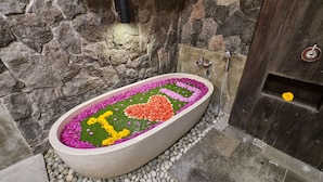 A stone-walled bathroom with a freestanding bathtub surrounded by pebbles and a floral arrangement. - Villa Casa Natura 15 (Ubud)