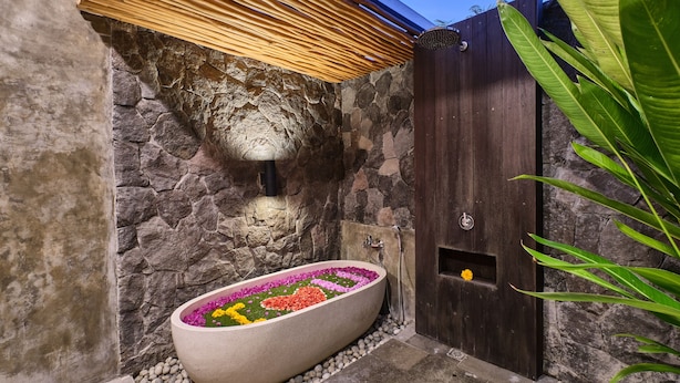A stone-walled bathroom with a freestanding bathtub filled with colorful flowers, a wooden door, and a stone wall. - Villa Casa Natura 15 (Ubud)