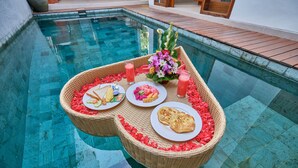 A poolside breakfast setup with a wicker tray containing plates of food, drinks, and a floral arrangement. - Villa Casa Natura 15 (Ubud)