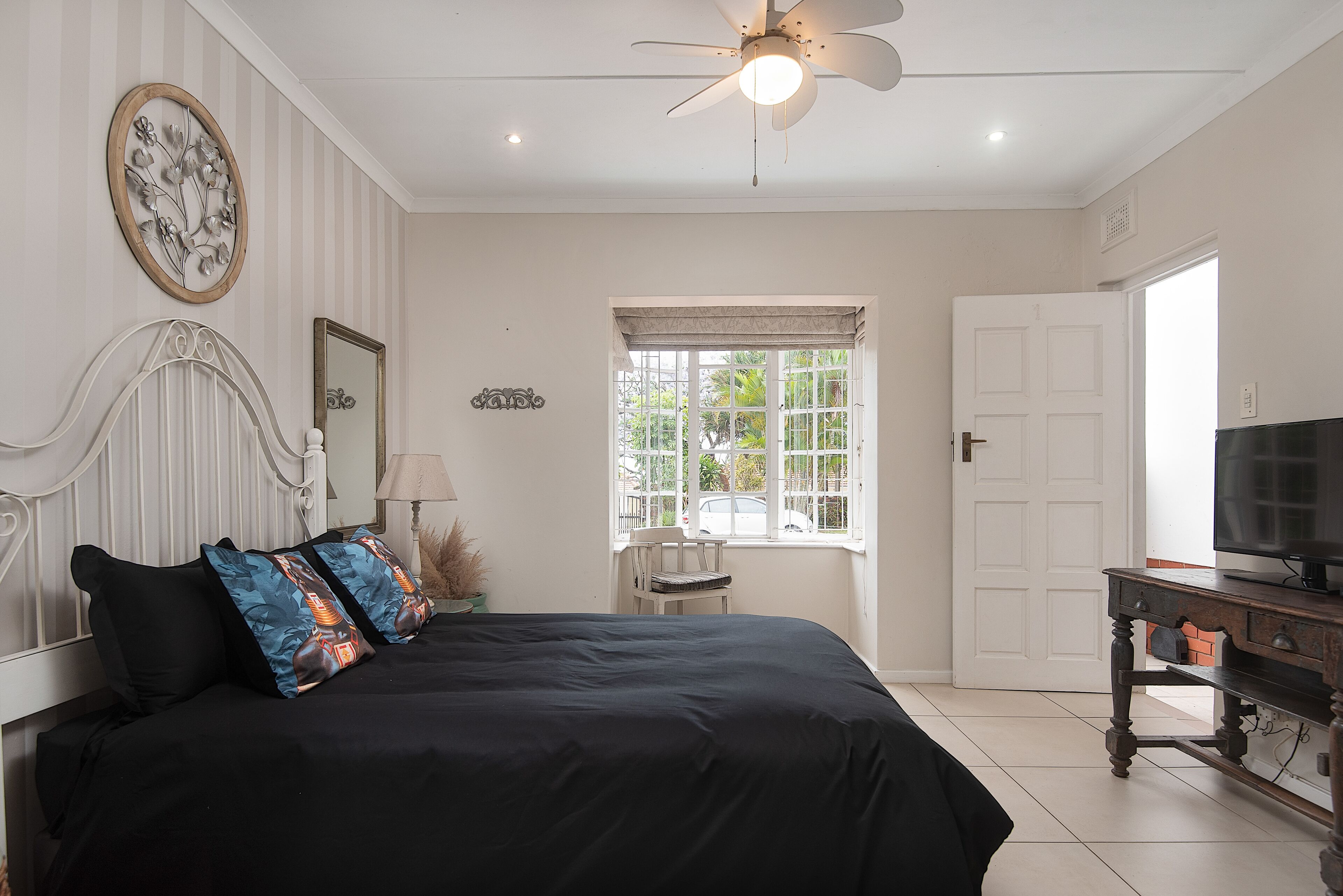 Un dormitorio con cama, ventilador de techo, ventana con vistas, puerta y televisor sobre un mueble.