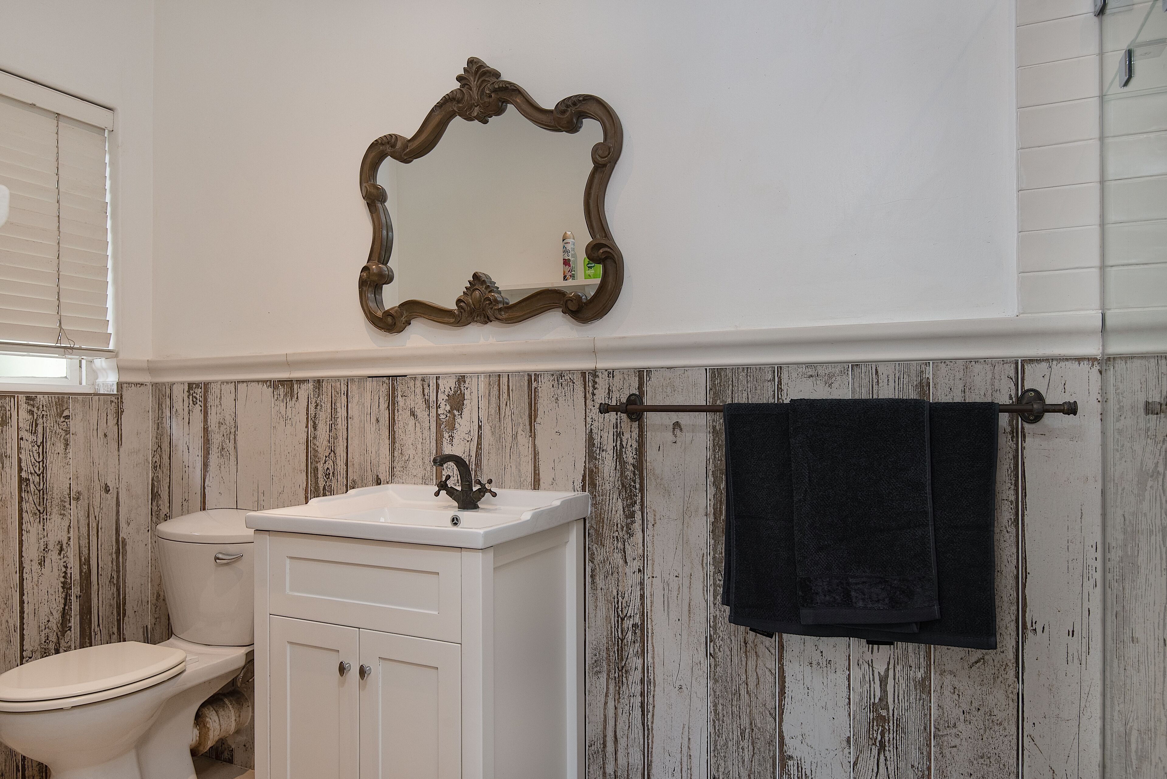 A bathroom with a white vanity, a toilet, a mirror with an ornate frame, and a towel rack with a black towel.