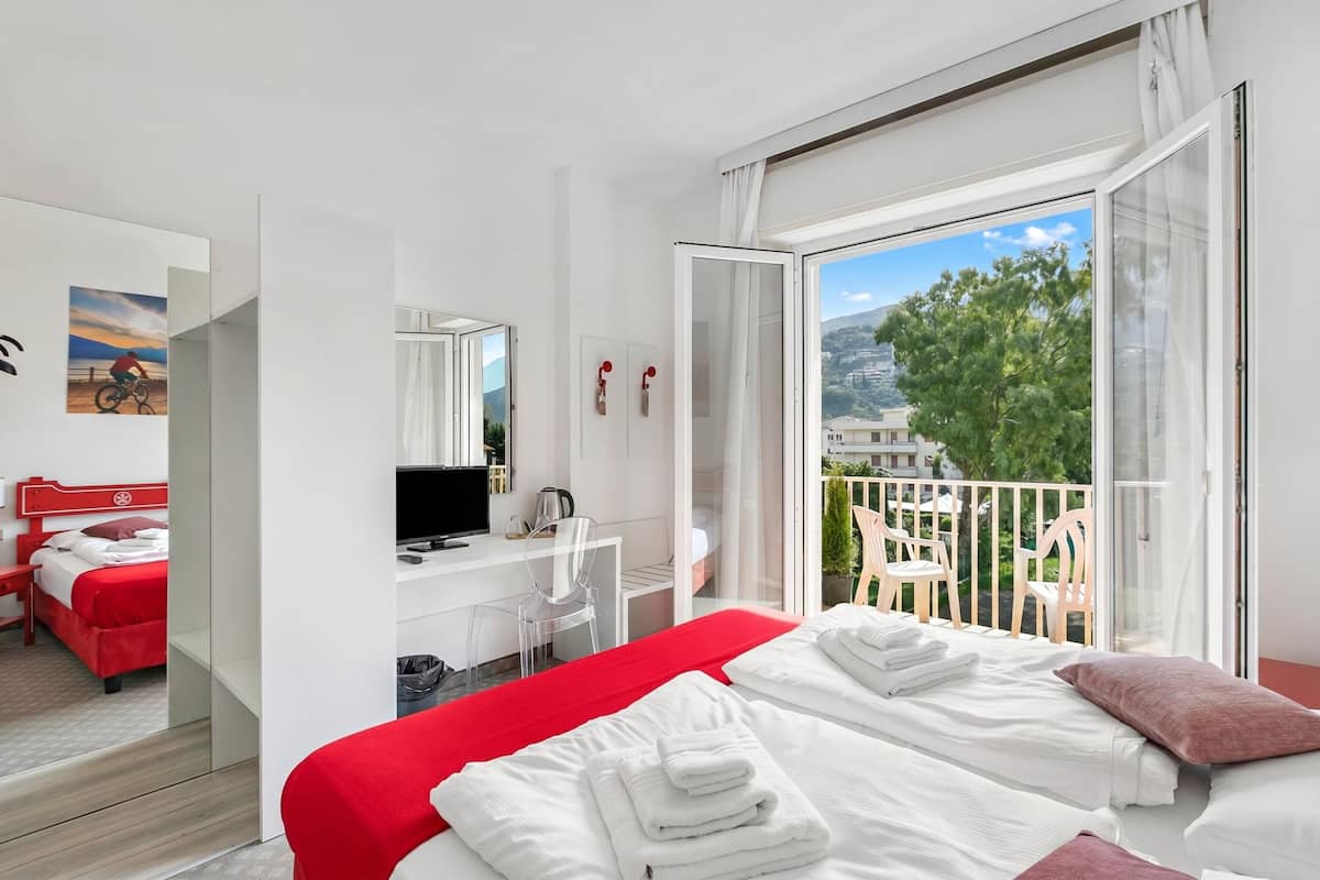A hotel room with a bed, a desk with a computer, a TV, a bathroom, and a balcony with chairs.