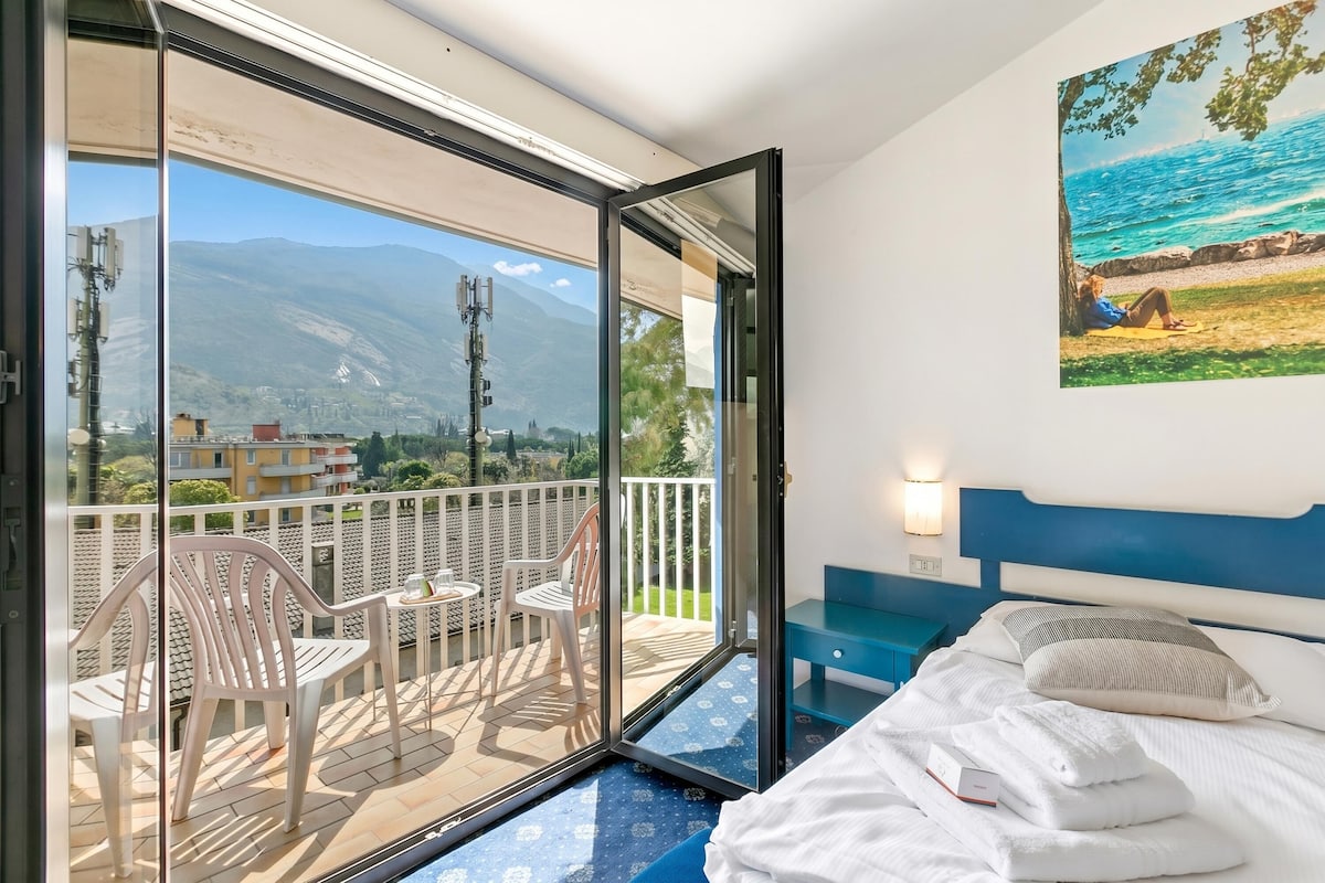 A hotel room with a balcony, a bed with white linens, a painting of a beach scene, and a view of mountains.
