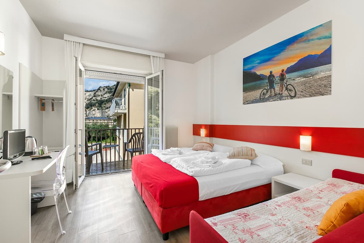 A hotel room with a bed, a desk, a chair, a balcony with a view of the mountains, and a painting of a sunset.