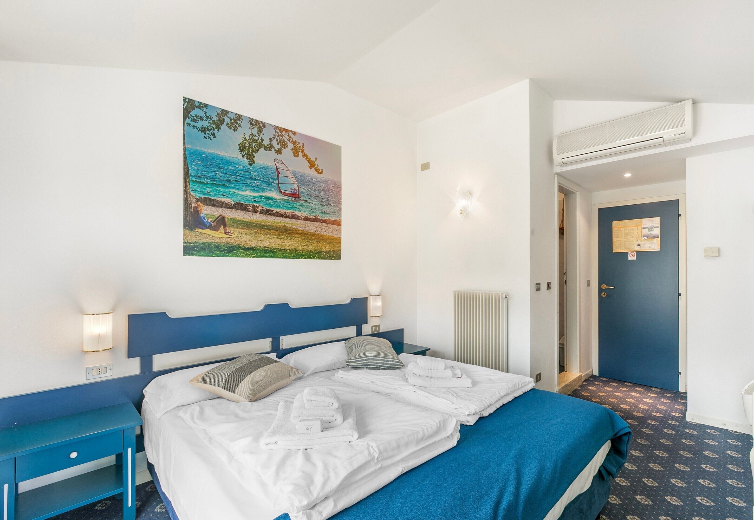 Een hotelkamer met een bed, nachtkastjes, een schilderij van een strandtafereel, een airconditioning en een blauwe deur.