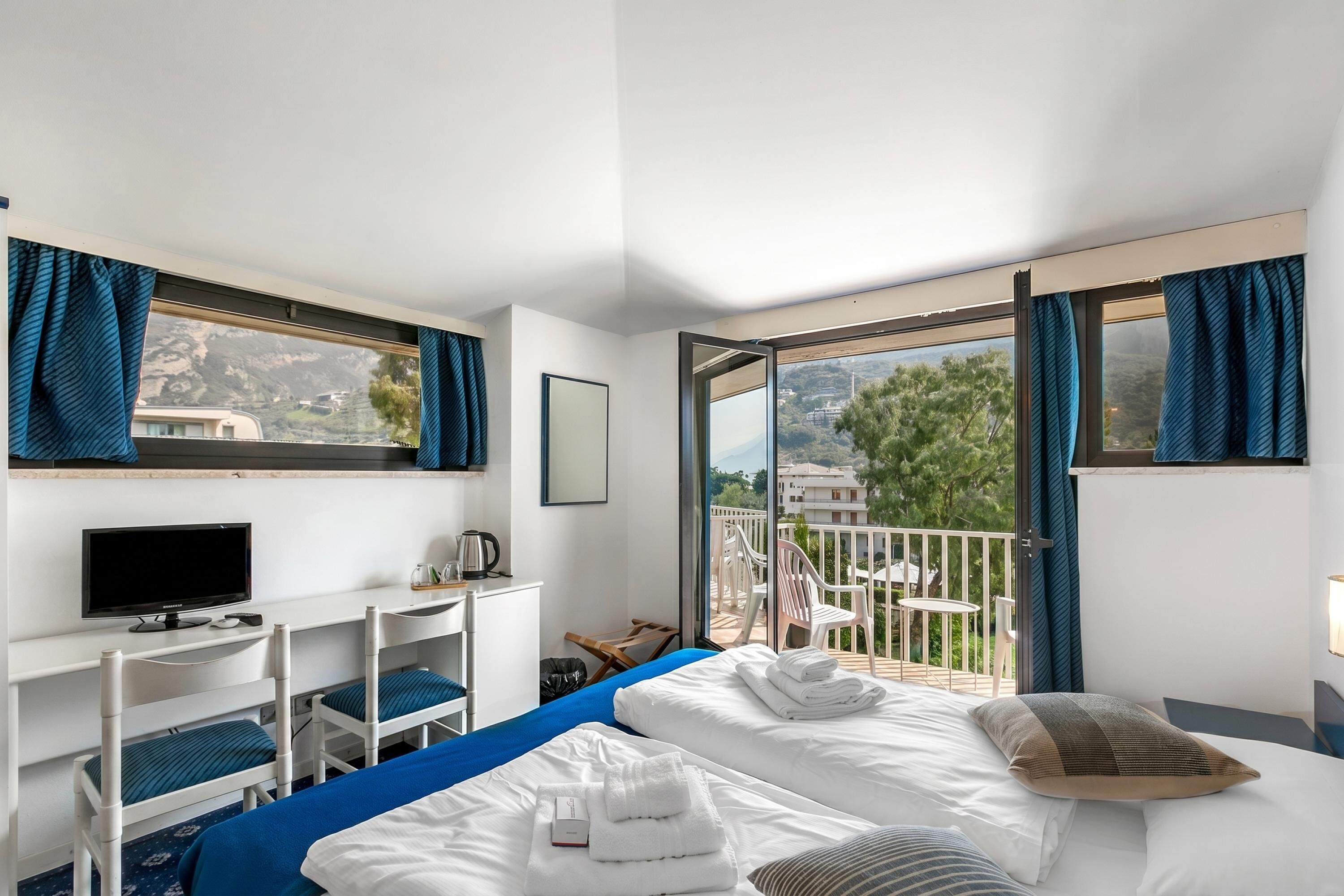 A hotel room with a bed, a desk with a TV, a balcony with chairs, and a view of the mountains.