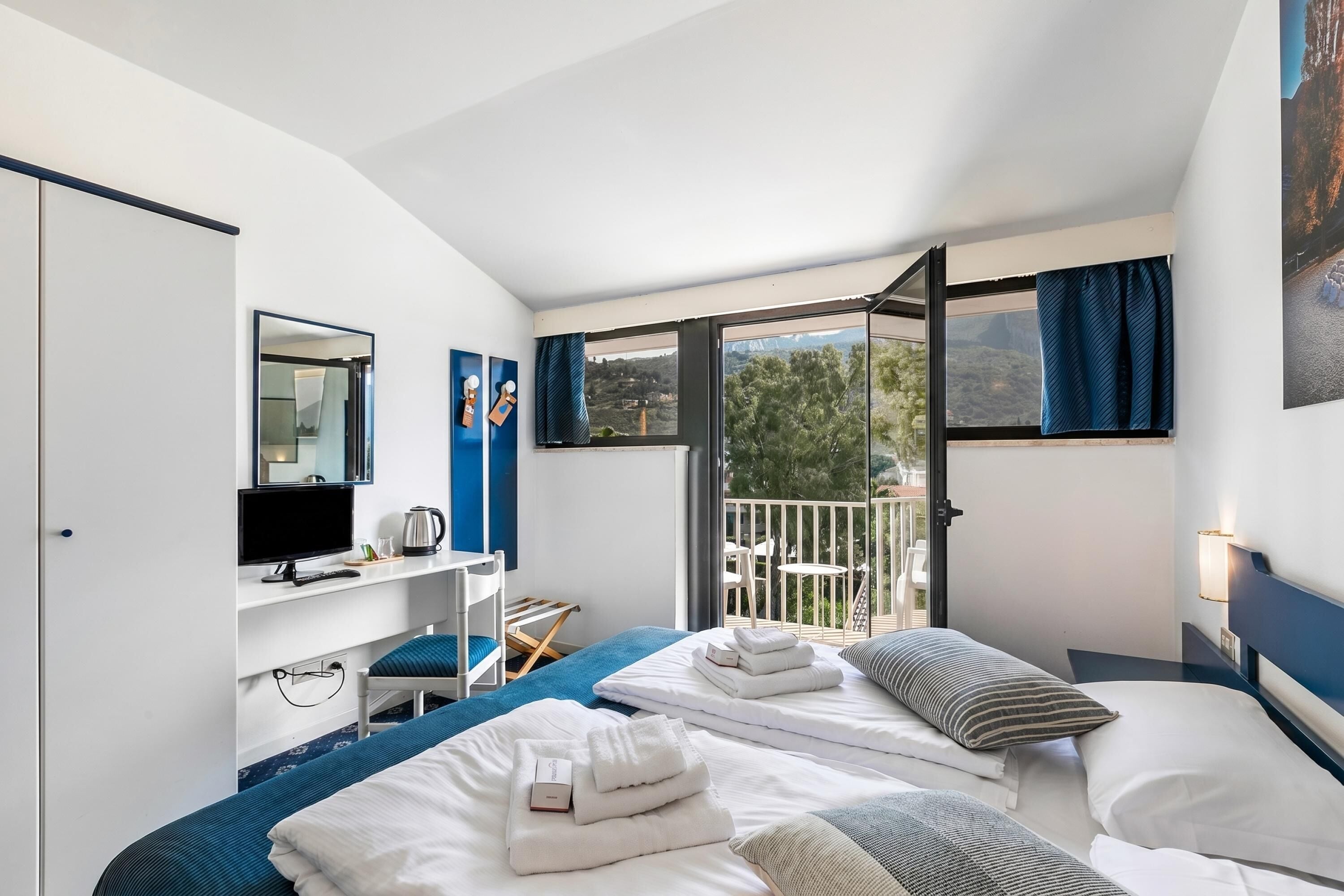 A hotel room with two beds, a desk with a TV, a chair, and a balcony with a view.