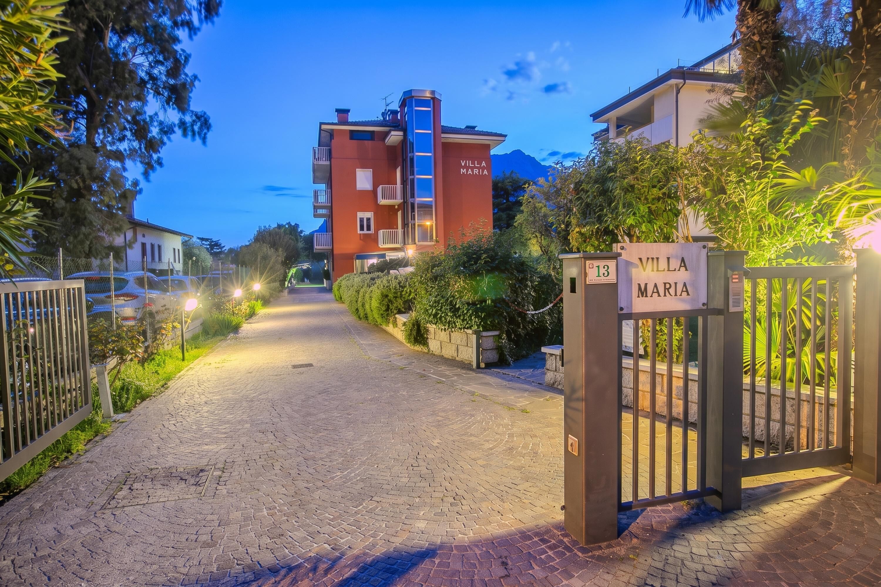 Ein gepflasterter Weg führt zur Villa Maria, umgeben von Grün und mit Blick auf die Berge.