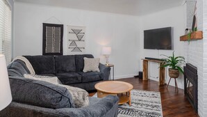 A modern living room with a dark grey sofa, a round wooden coffee table, a TV mounted on the wall, and a fireplace. - Charming Downtown Bungalow with Yard & Gas Grill (Oshkosh)
