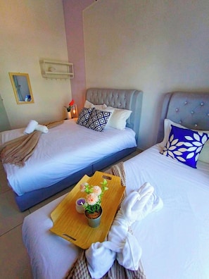 A hotel room with two beds, each with a tufted headboard, white linens, and decorative pillows. A wooden tray with flowers and a candle is placed on the bed.