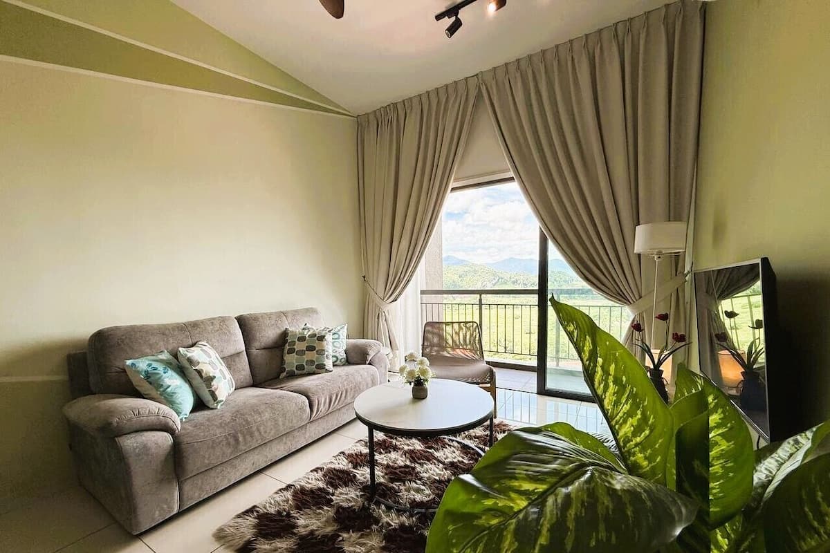 Un salon avec un canapé, une table basse ronde, une grande plante et un balcon avec des rideaux.
