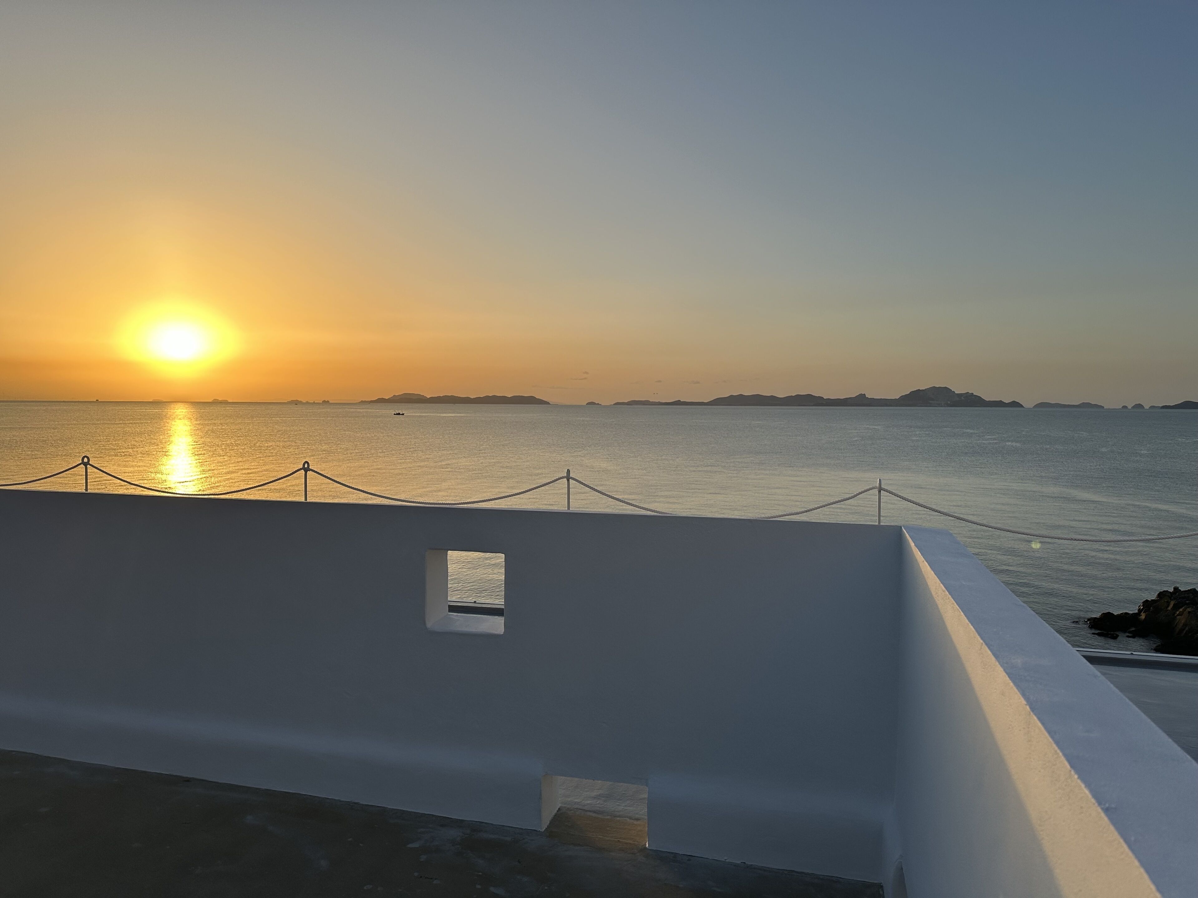 Vista al mar con un atardecer, un edificio blanco con una pequeña ventana y una barandilla de cuerda.