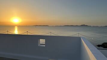 Vista al mar con un atardecer, un edificio blanco con una pequeña ventana y una barandilla de cuerda.