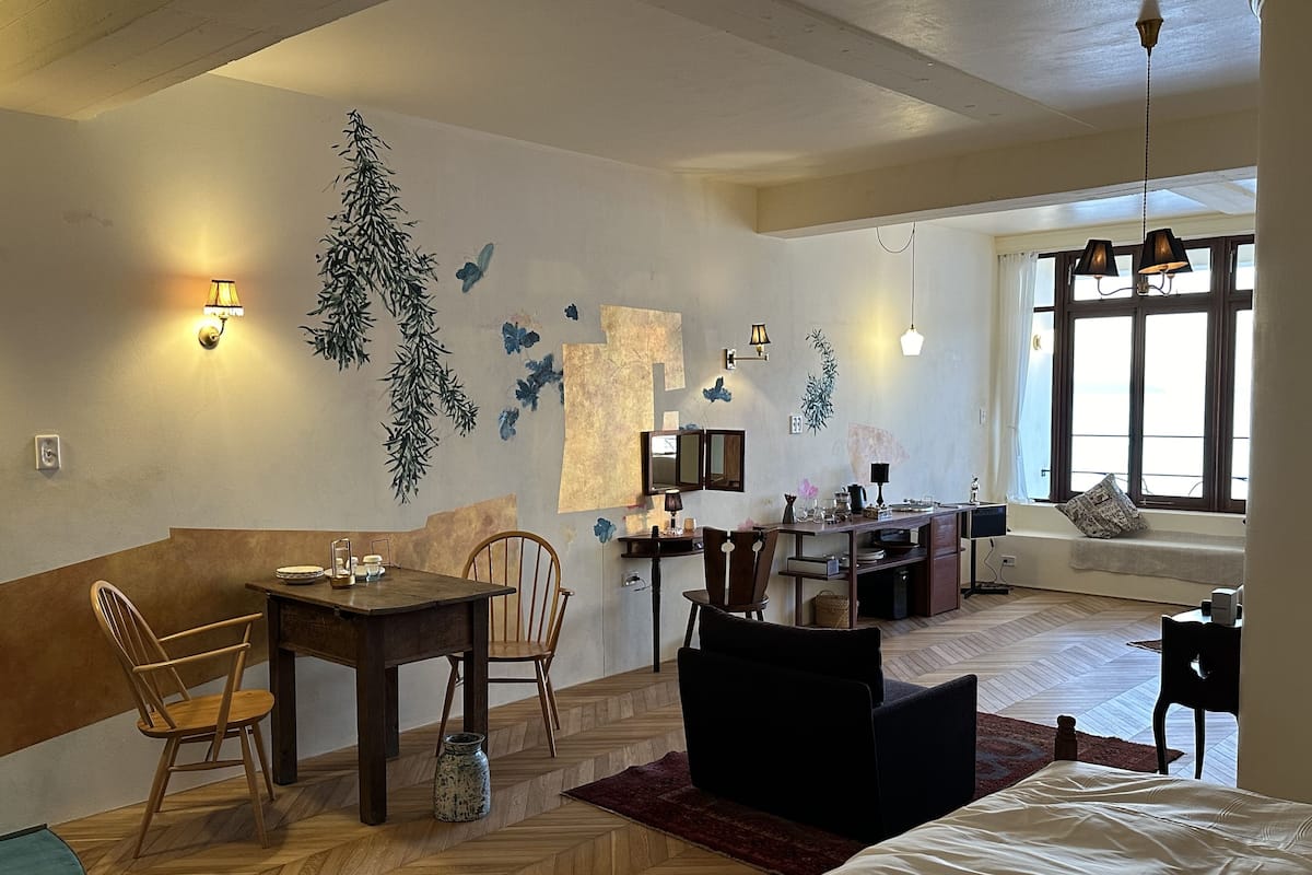 Ein gemütliches Wohnzimmer mit Holzdielen im Fischgrätmuster, einer Couch, einem Esstisch mit Stühlen und einer Küchenzeile mit Fenster.