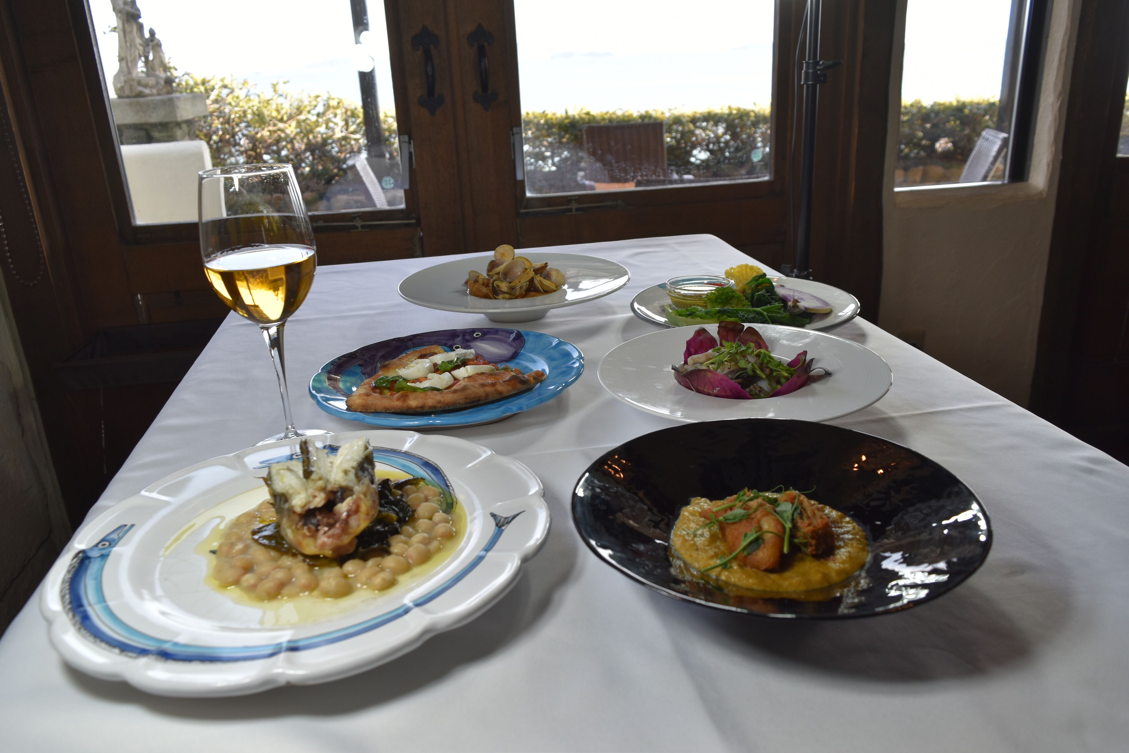 A table set with various dishes and a glass of white wine, with a view of greenery outside.