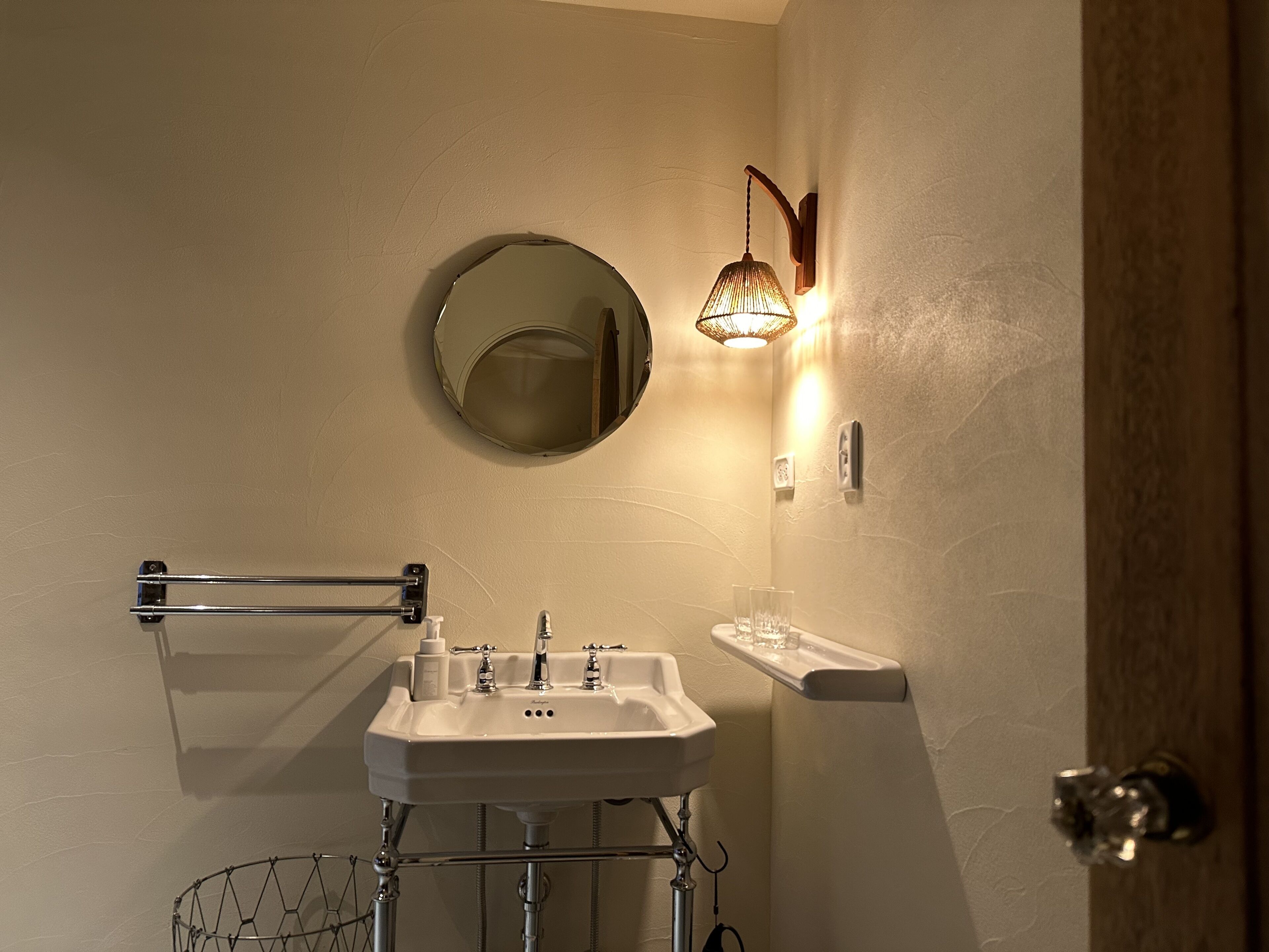 A bathroom with a white sink, a round mirror, a towel rack, a glass shower enclosure, and a wall-mounted light fixture.
