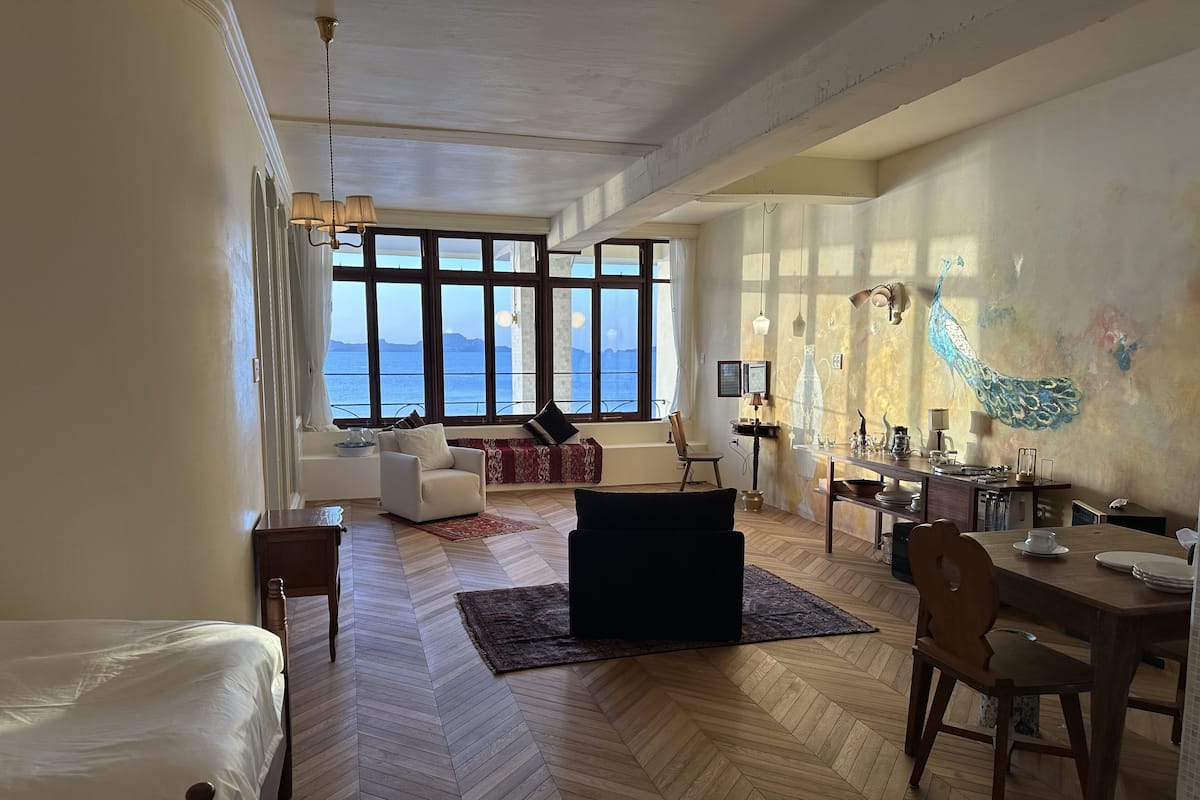 Ein geräumiges Wohnzimmer mit großem Fenster zum Meer, Holzboden und einem Essbereich mit Tisch und Stühlen.
