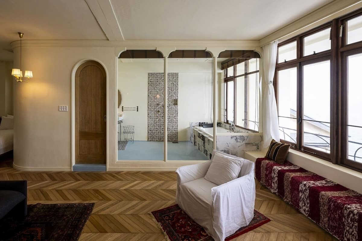 Ein Hotelzimmer mit einem großen Bett, einem Schlafsofa, einem Sessel, einem Spiegel und Blick auf die Stadt durch große Fenster.