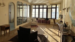Amplia sala de estar con ventanas grandes, piso de madera en espiga, un sofá, una silla y una mesita con una lámpara.