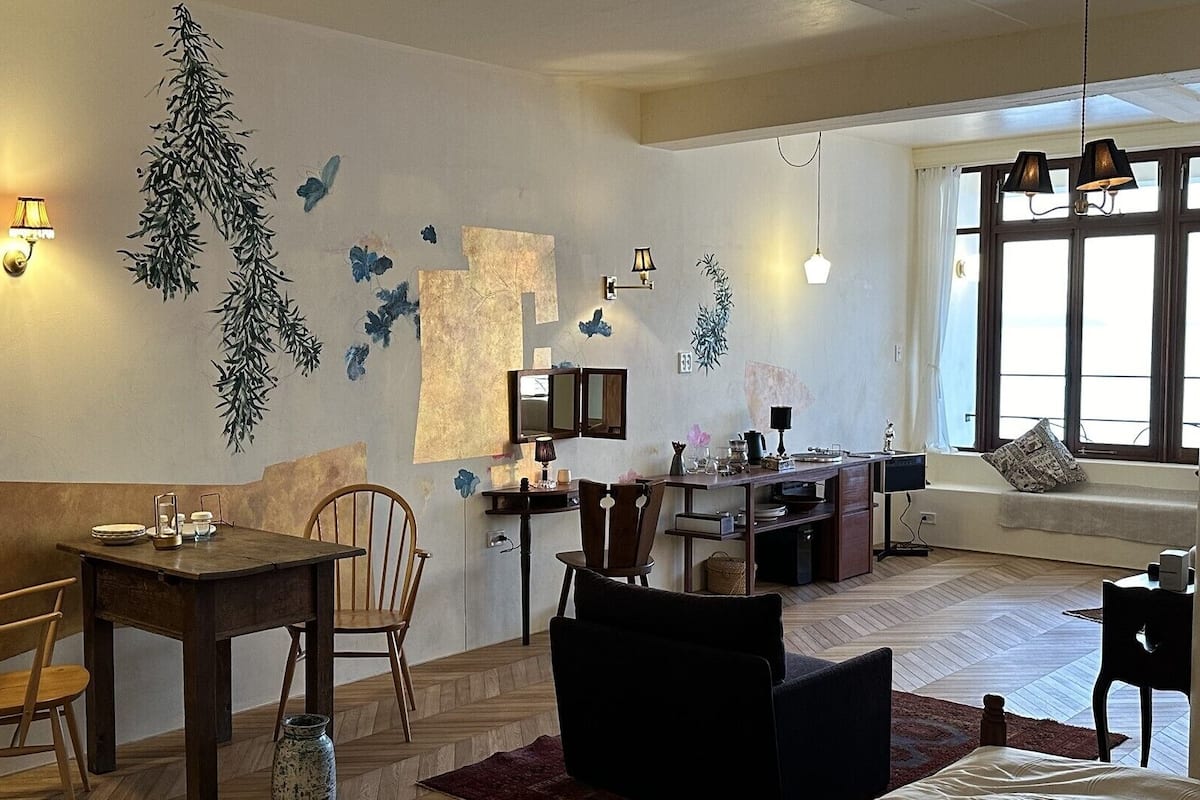Ein Essbereich mit Holztisch und Stühlen, eine Küchenzeile mit Arbeitsplatte und Geräten sowie eine Sitzecke mit Sofa und einem Vorhangfenster.