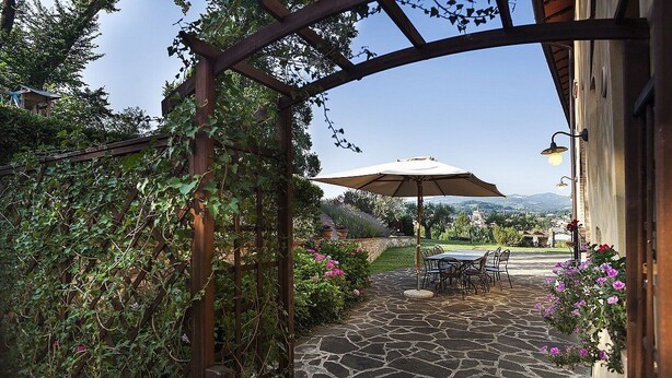 A stone pathway leading to an outdoor seating area with a pergola and umbrella, surrounded by lush greenery and a view of distant hills. - Casa Gioele D: A welcoming apartment situated in a quiet location, a few minutes from the town center, with Free WI-FI. (Umbertide)