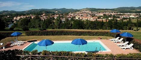 Una piscina con sillas de descanso y sombrillas, rodeada por una valla de madera, con vista a un pueblo al fondo.