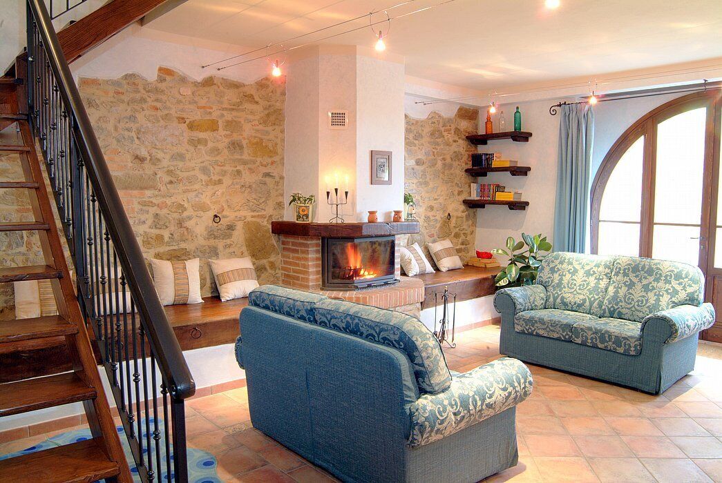 A cozy living room with a fireplace, stone walls, a wooden staircase, and a large window.