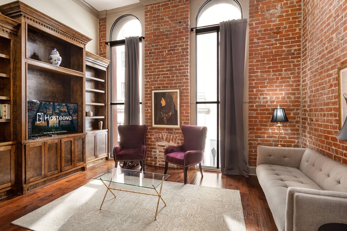 A living room with a brick wall, a sofa, two armchairs, a glass coffee table, a TV, and a bookshelf.