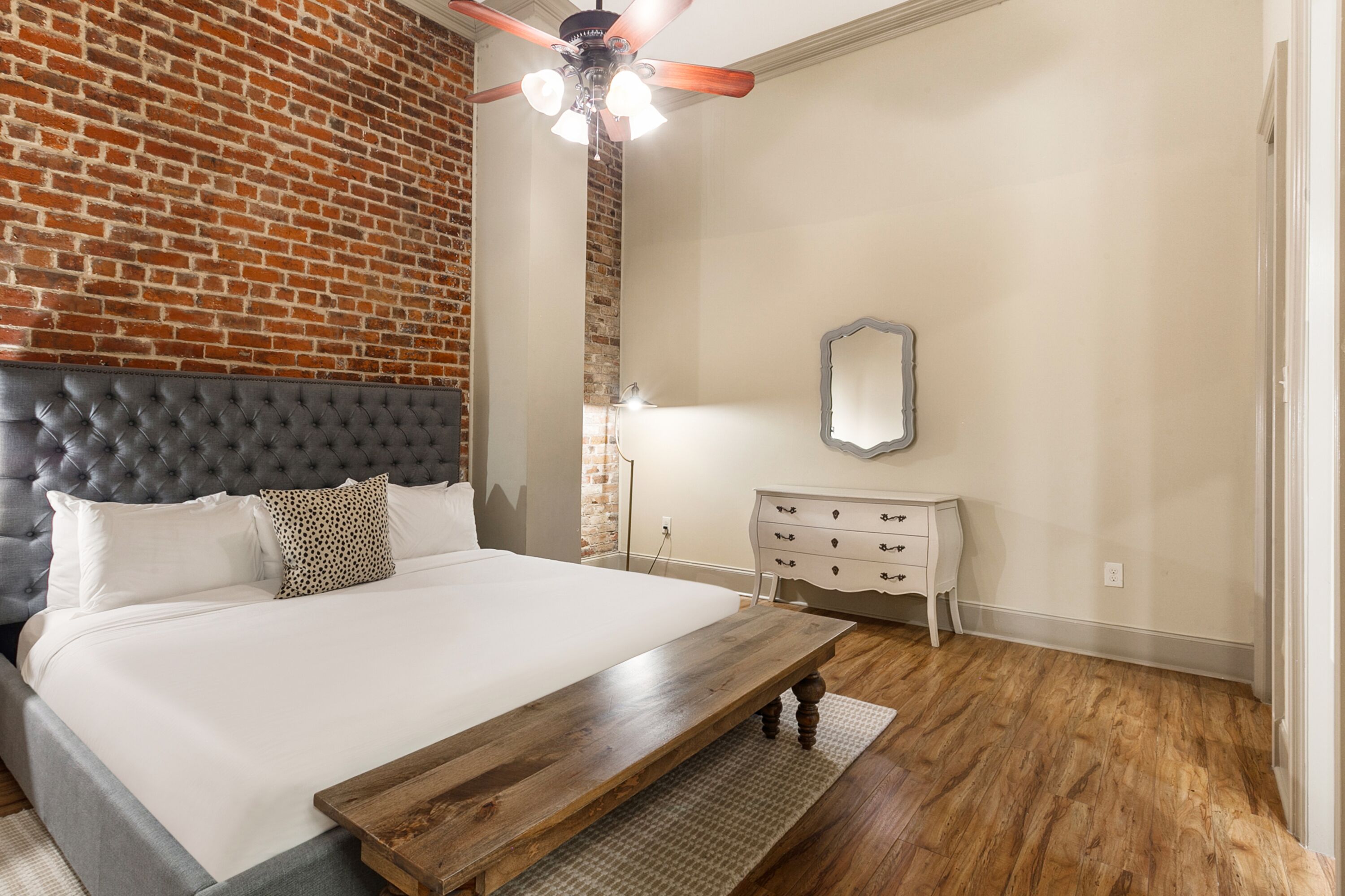 Un dormitorio con pared de ladrillo, cabecera acolchada, estructura de cama de madera, mesita de noche, espejo y ventilador de techo.