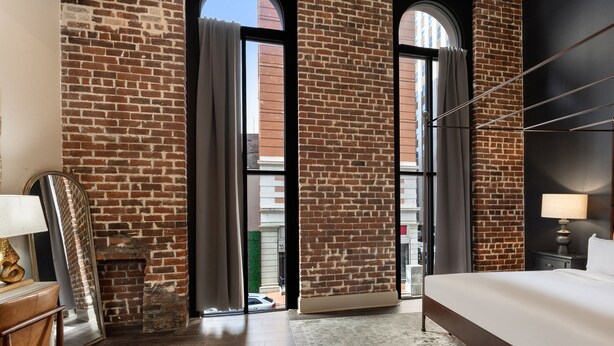A hotel room with a brick wall, a large bed, a desk with a lamp, and a view through a glass door. - Chuck's place (New Orleans)