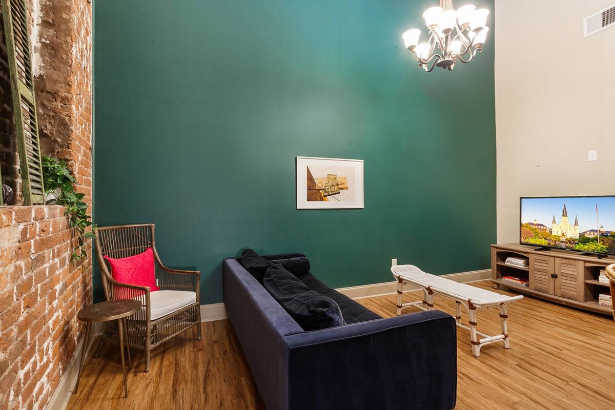 A cozy living room with a brick wall, a dark green wall, a wooden bench, a wicker chair, a sofa, and a television.