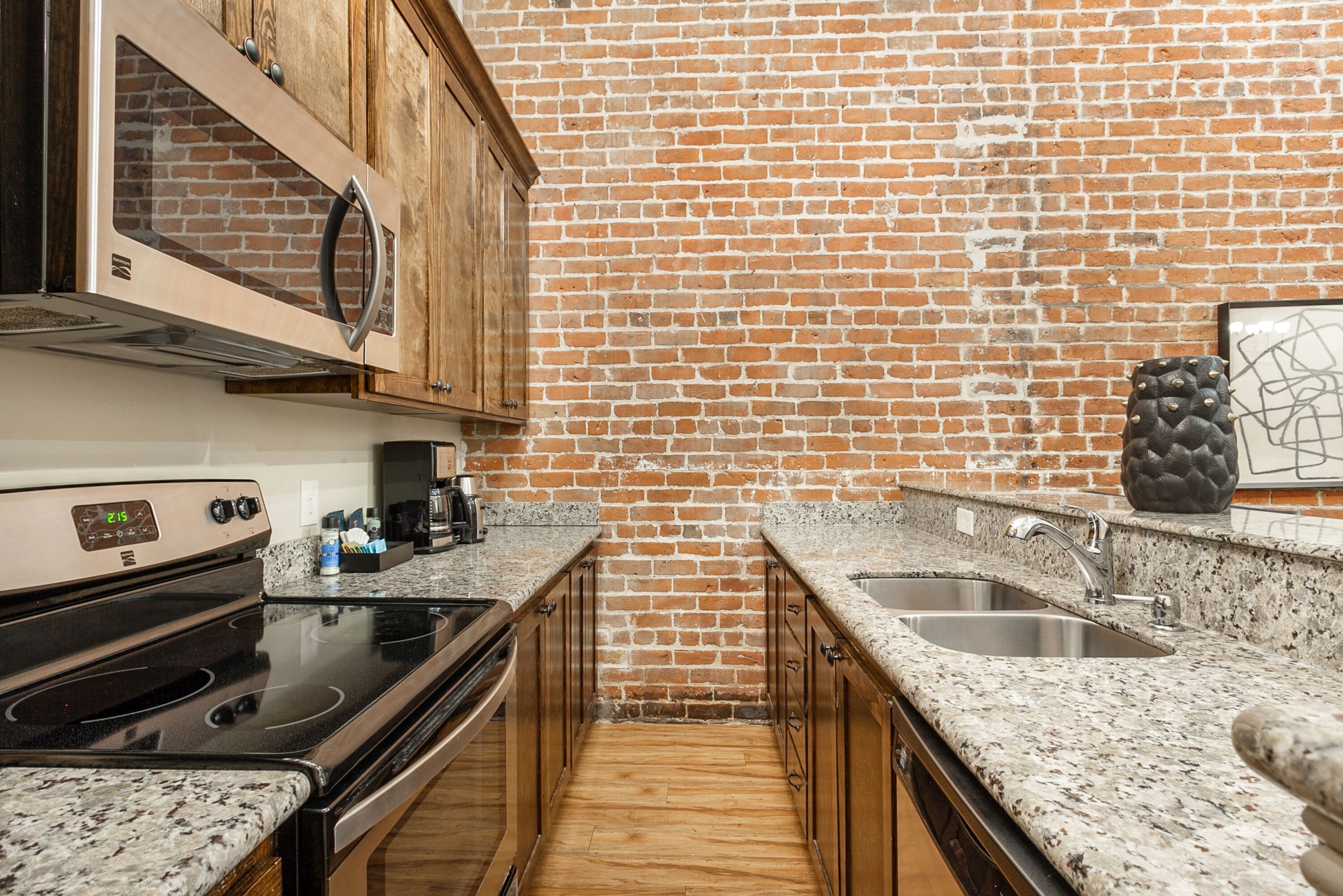 A kitchen with a brick wall, wooden cabinets, a stainless steel sink, and a granite countertop.