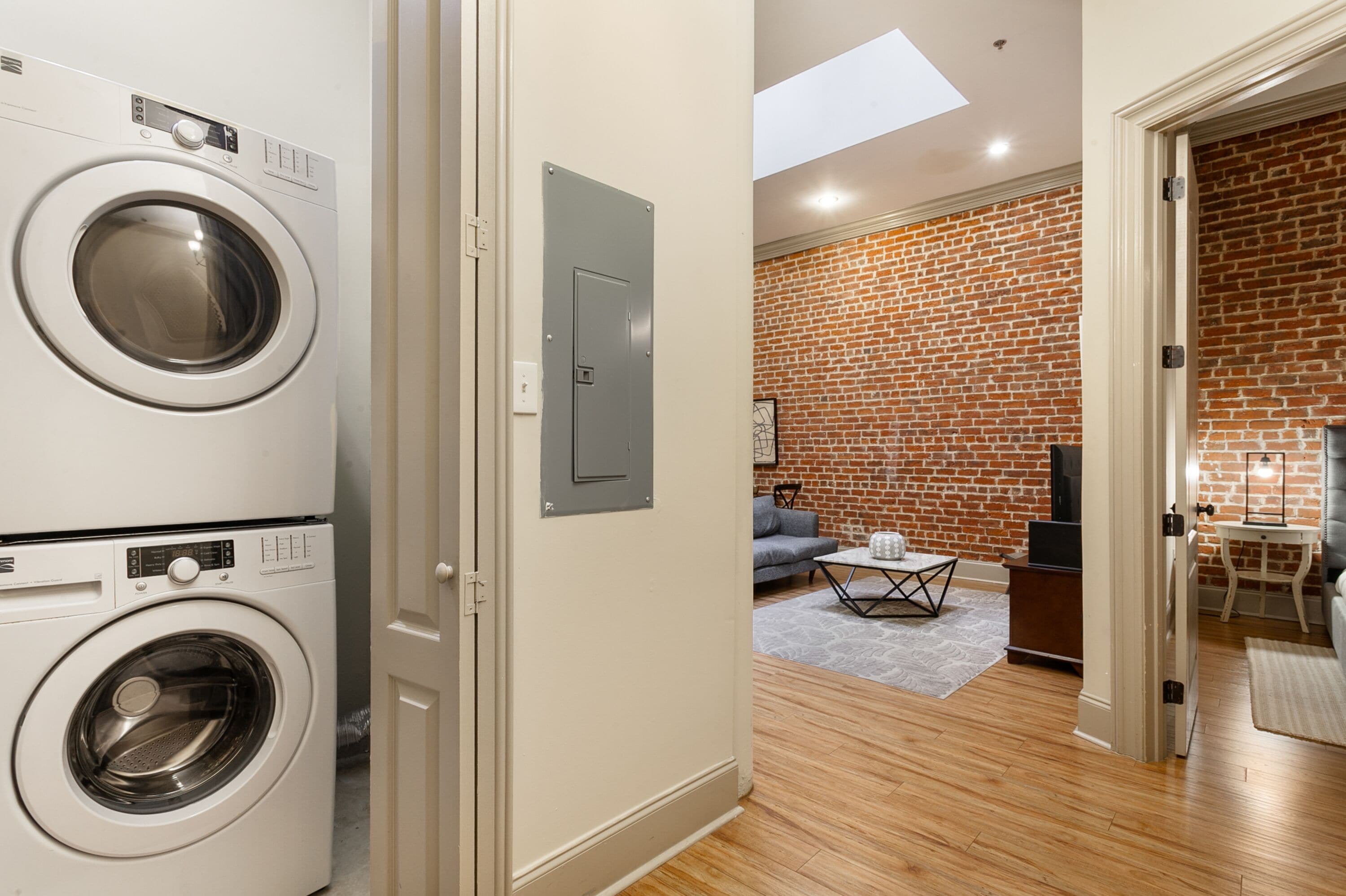A laundry room with a front-loading washer and dryer, a brick-patterned living room with a sofa and coffee table, and a skylight above the laundry area.