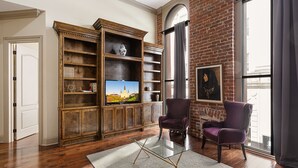 A living room with a brick wall, wooden shelving, a television, two armchairs, and a glass coffee table. - Chuck's place (New Orleans)