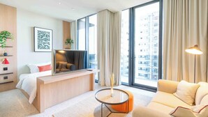A modern hotel room with a bed, a desk, a sofa, a TV, and large windows offering a city view. - Prime Location, Pristine Studio in Business Bay (Dubai)