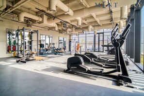 Une salle de sport équipée de matériel d’exercice, dotée de grandes fenêtres et d’un plafond haut avec des conduits apparents.