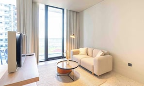 A modern hotel room with a sofa, a coffee table, a TV, and a large window offering a view of the city.