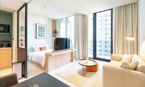 A modern hotel room with a bed, a TV, a sofa, a small table, and large windows offering a city view.