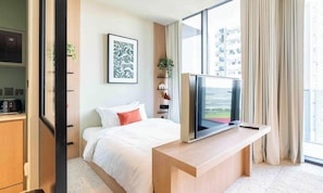A modern hotel room with a bed, a desk with a TV, a window with curtains, and a city view.