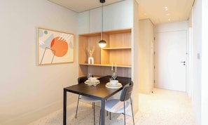 A modern dining area with a dark wooden table, two chairs, and a pendant light. A framed abstract artwork is on the wall, and there is a wooden shelf with decorative items.