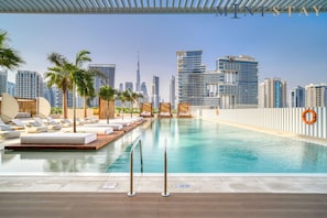 A rooftop infinity pool with lounge chairs and palm trees, overlooking a city skyline with modern buildings.