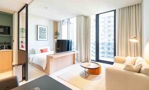 Moderni hotellihuone, jossa on sänky, televisio, sohva, pieni pöytä ja suuret ikkunat, joista avautuu näkymä kaupunkiin.