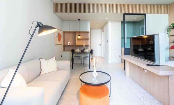 Ein modernes Wohnzimmer mit einer Couch, einem Couchtisch und einer Stehlampe. Im Hintergrund ist ein Essbereich mit Tisch und Stühlen zu sehen.