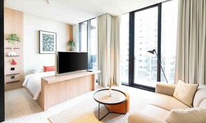 A modern hotel room with a large window, a flat-screen TV, a sofa, a coffee table, and a bed with white bedding.