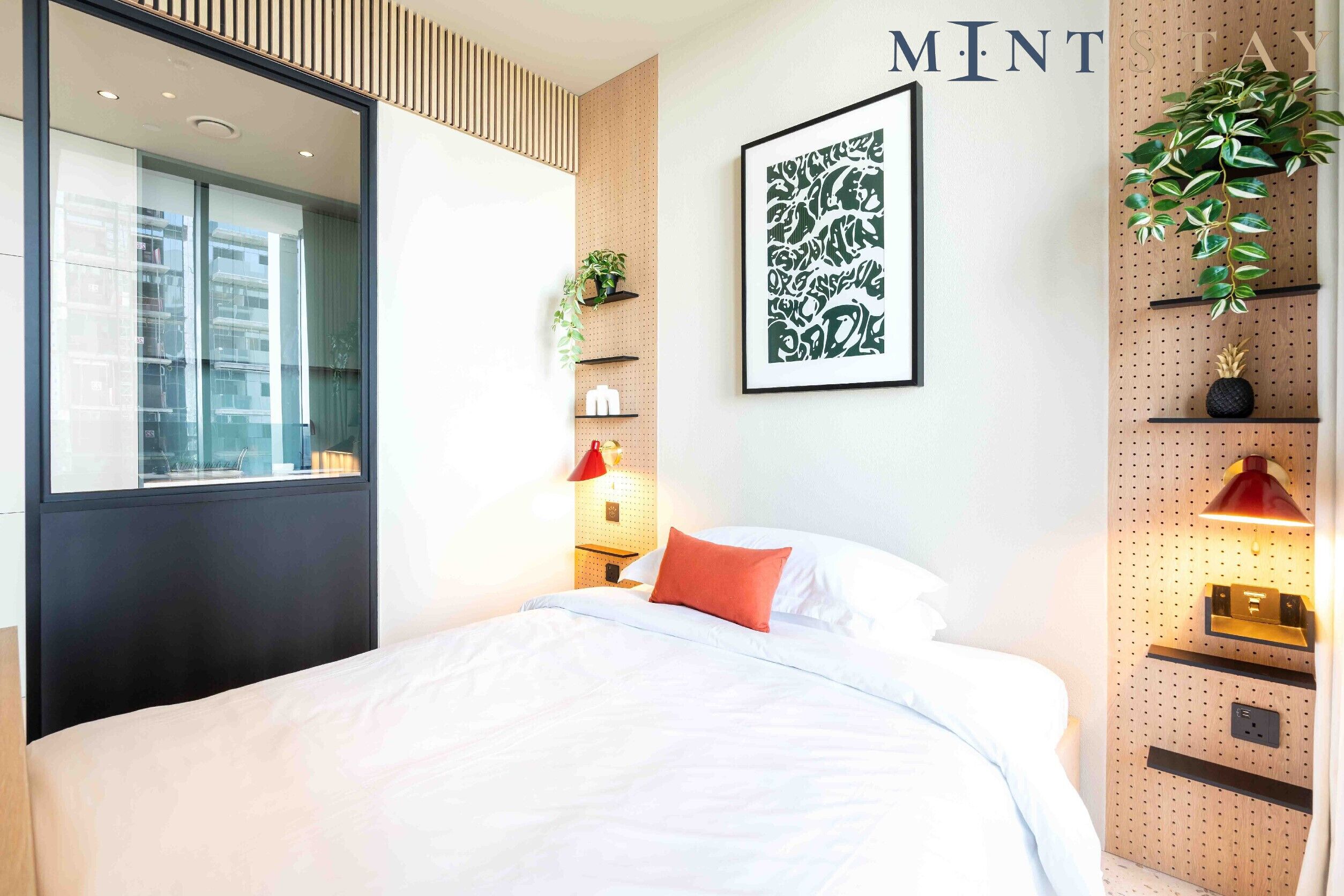 Une chambre d’hôtel moderne avec un lit, une étagère murale agrémentée de plantes, une œuvre d’art encadrée et un panneau indiquant « MINT STAY ».