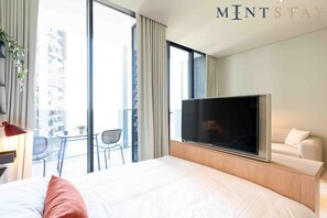 A hotel room with a large bed, a TV, a balcony with a table and chairs, and a view of a building.